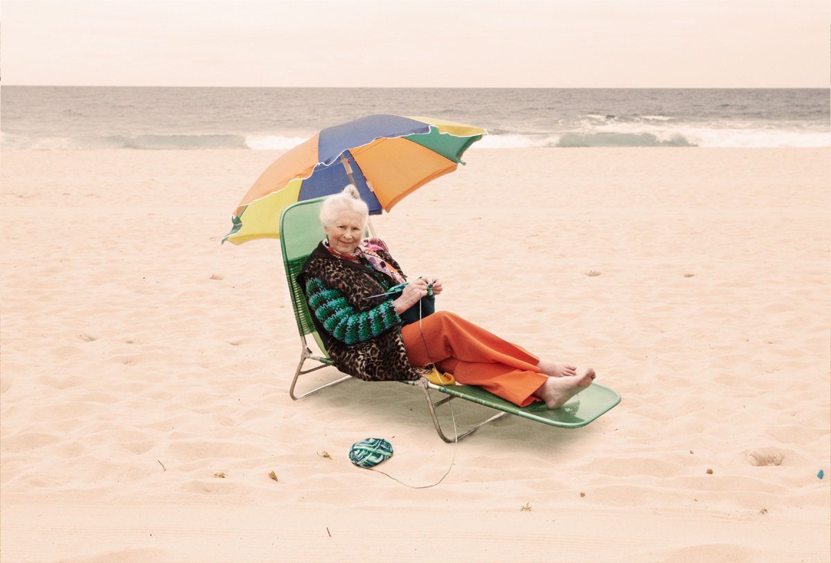 Elderly woman relaxes in a beach chair under a colorful umbrella, knitting with green yarn, the ocean visible in the background.