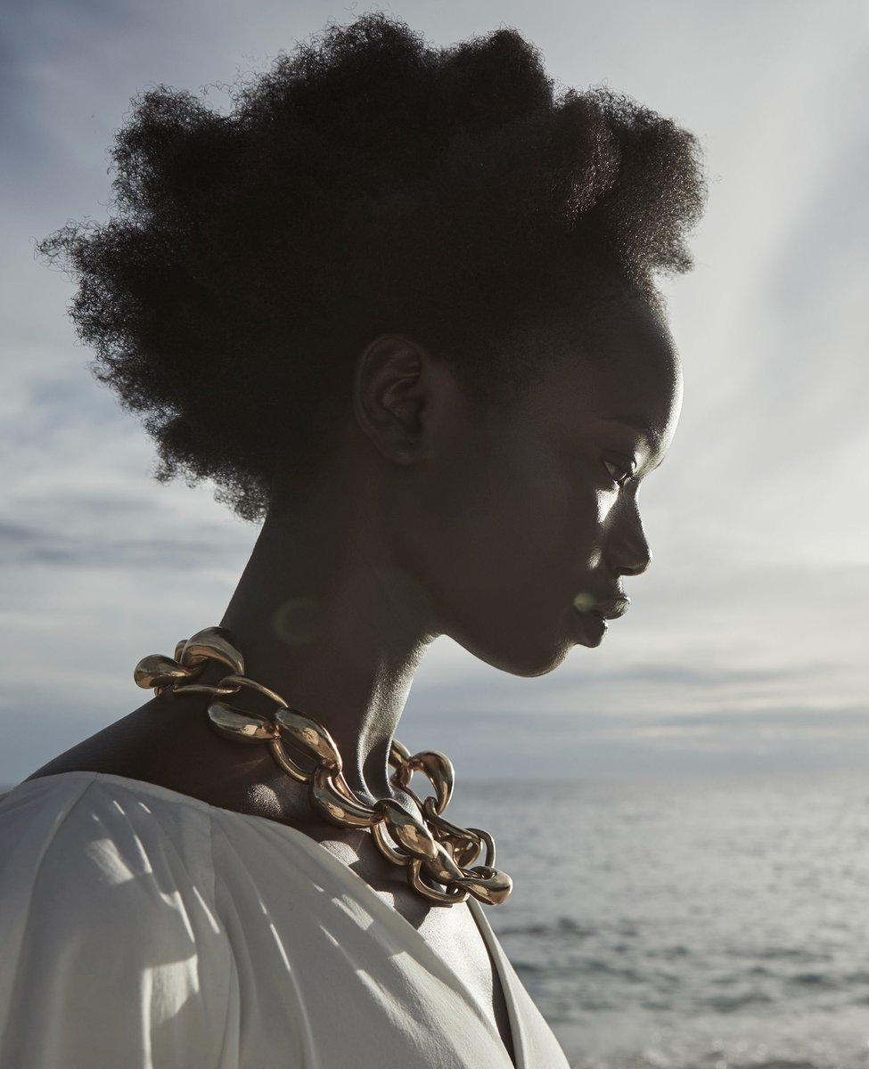 Profile shot of a dark-skinned woman with natural hair, wearing a white top and a large gold necklace, against a soft, light sky background.