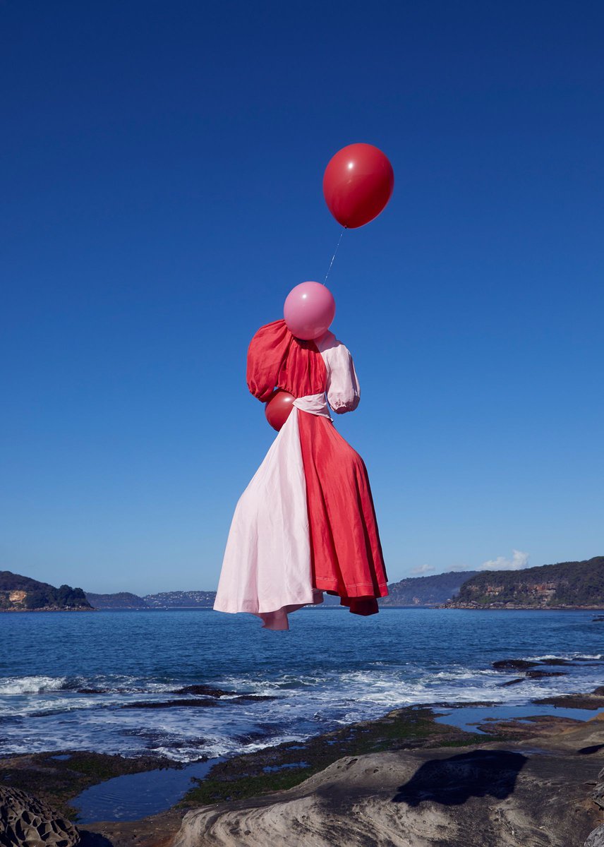 Levitating woman in a pink and red dress holds pink and red balloons against a blue sky backdrop above a rocky beach shoreline.