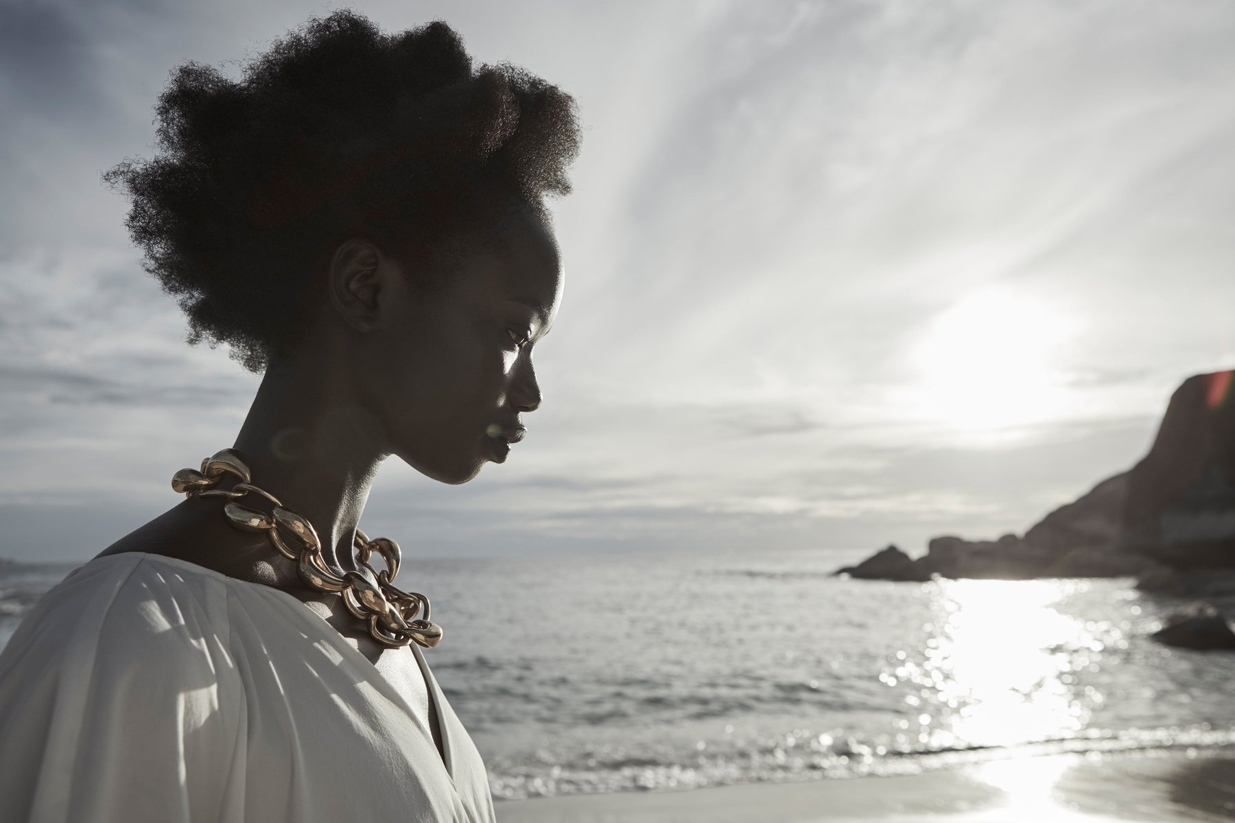 Black woman in profile wearing a white top and gold chain necklace by a beach at sunset, photographed by Juli Balla.