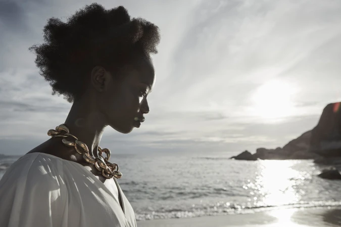 Black woman in profile wearing a white top and gold chain necklace by a beach at sunset, photographed by Juli Balla.