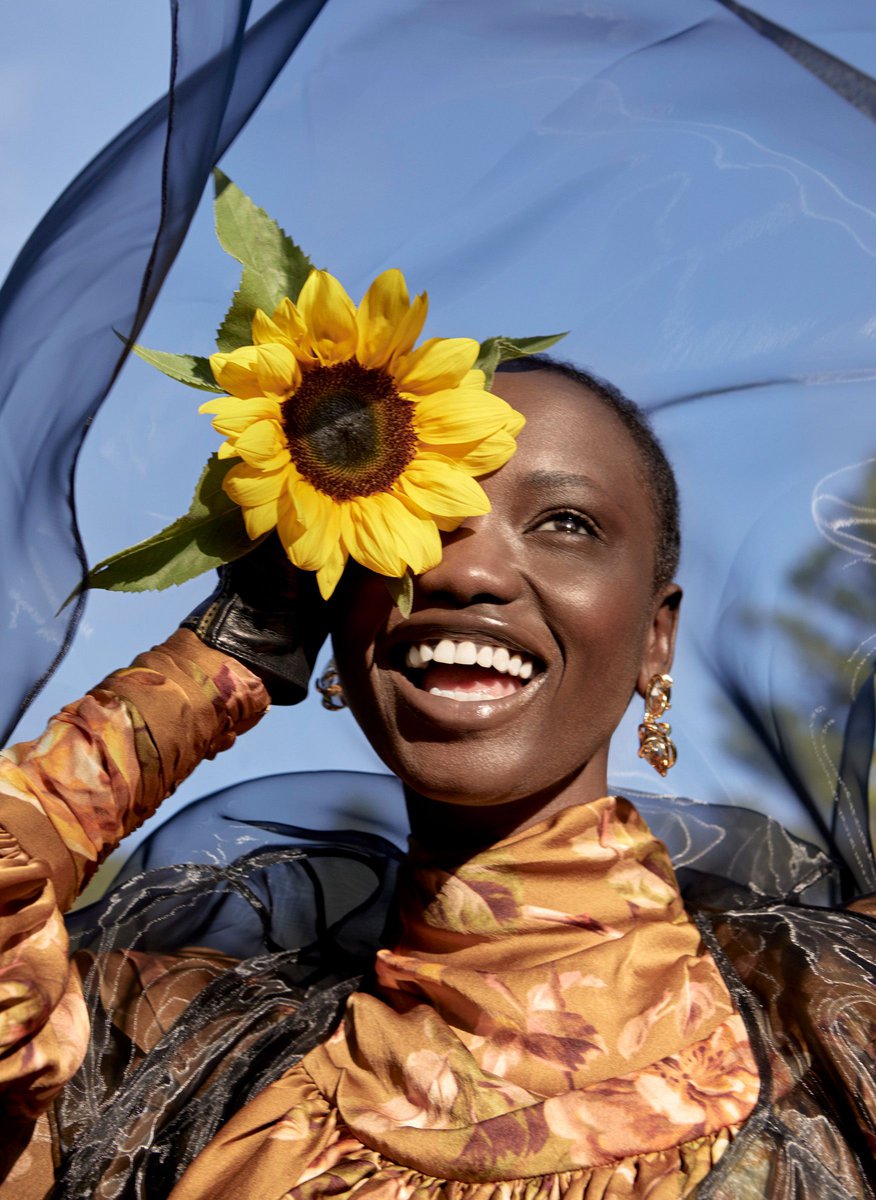 Smiling Black woman in patterned dress wearing a sunflower against a blue sky, accented by a sheer black shawl. Bright, fashionable.