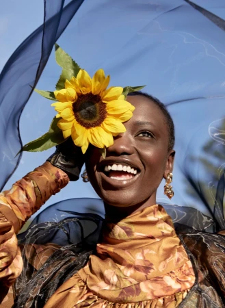 Smiling Black woman in patterned dress wearing a sunflower against a blue sky, accented by a sheer black shawl. Bright, fashionable.