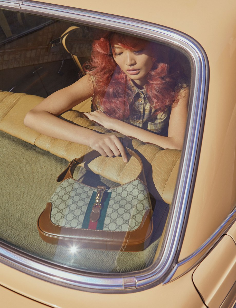 Woman with wavy hair and tan complexion inside a vintage car, posing with a patterned handbag; filtered sunlight and golden tones.