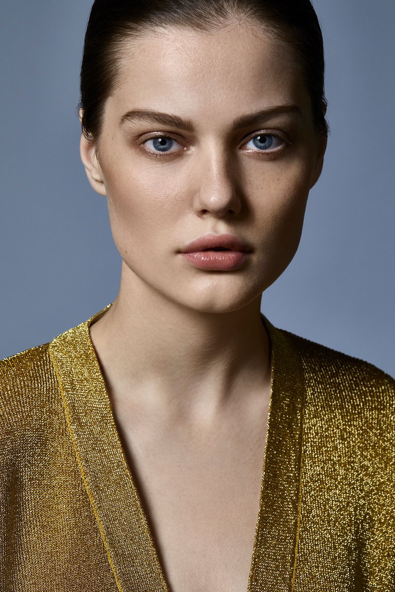 Close-up portrait of an adult female model with fair skin, blue eyes, and slicked-back hair, wearing a gold shimmer top, against a light blue background.