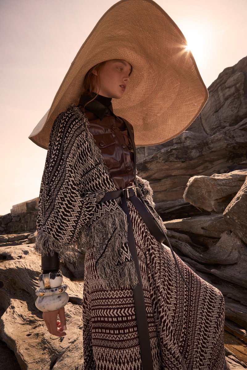 Woman in a large straw hat, patterned poncho, and maxi skirt poses outdoors against a rocky background in bright, diffused sunlight.