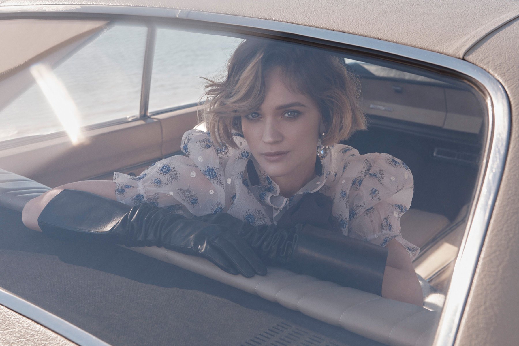 A woman with short brown hair and a floral top gazes out the back window of a classic car, wearing long black gloves in soft light.
