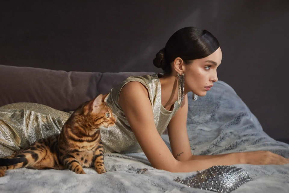 Woman in gold dress poses with a Bengal cat on a fur-covered bed, showcasing fashion and feline companionship in a soft, warm indoor setting.