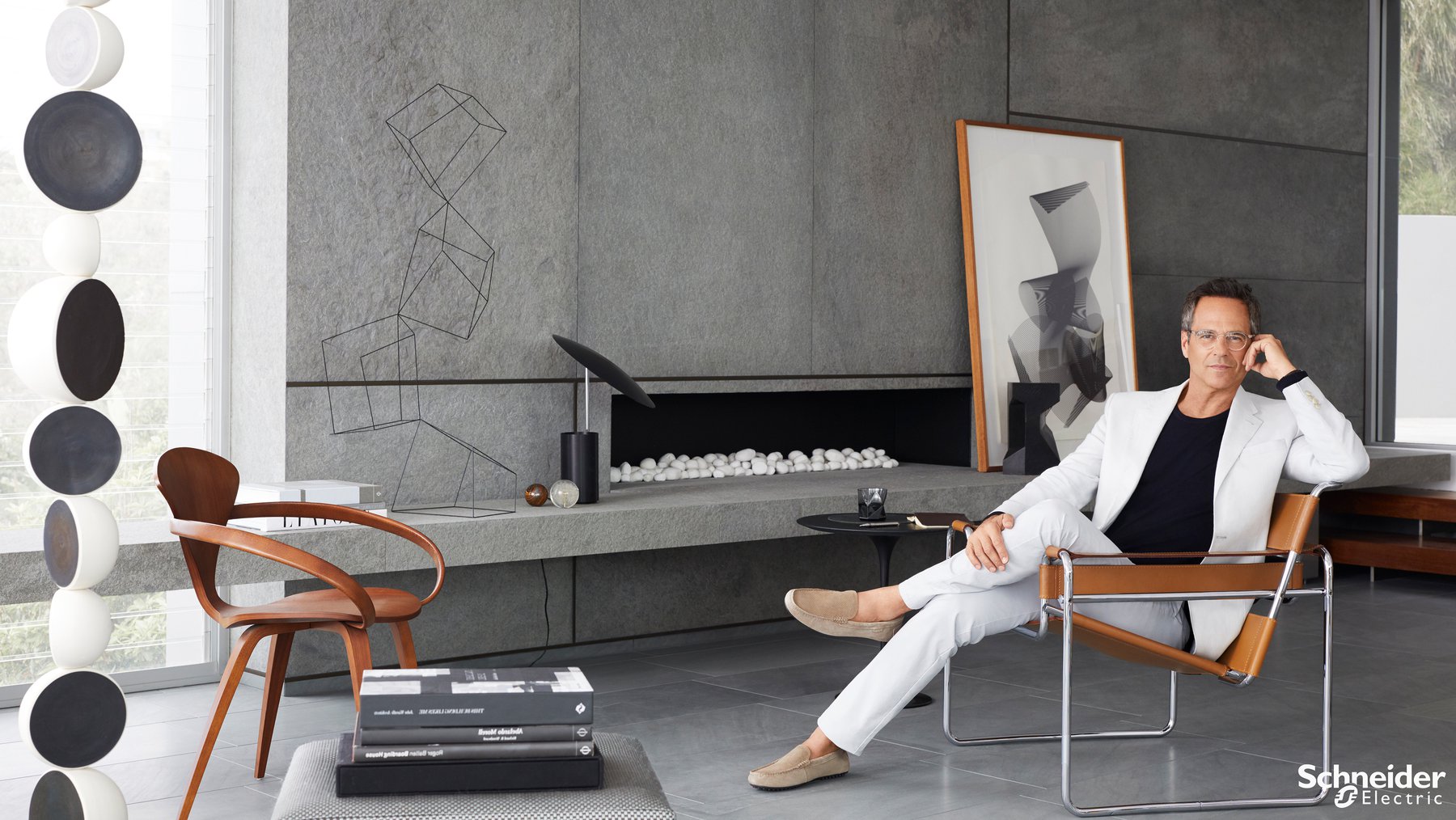 Modern interior view showcases a seated man in white suit, alongside geometric art, stylish furniture, and natural light from a window.