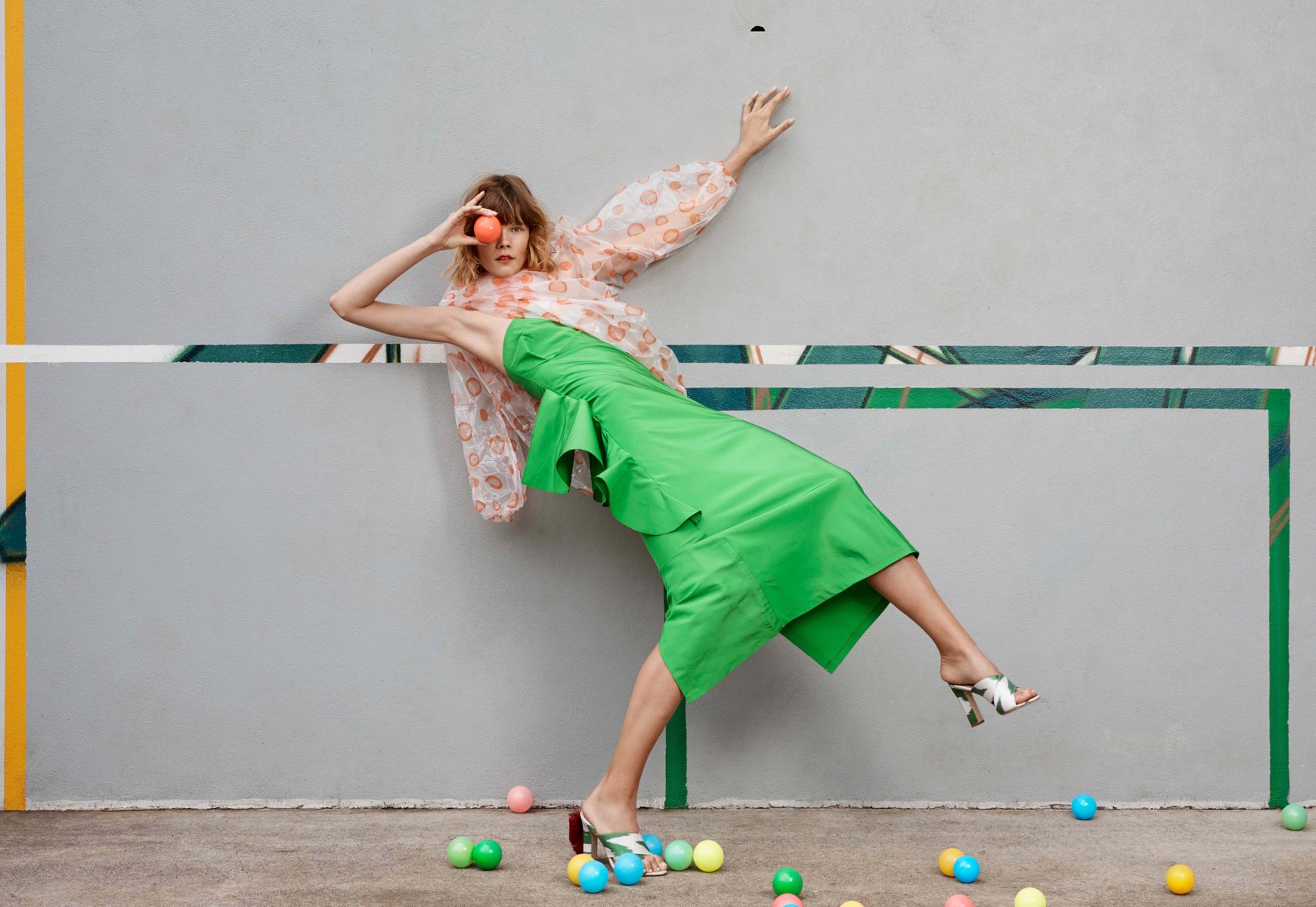 Woman strikes a pose in a green dress and patterned top, balancing an orange ball in front of her eye near colorful balls on the ground.