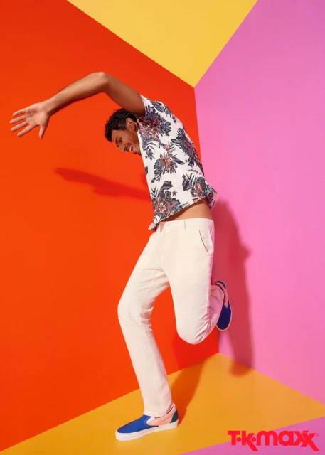 Man strikes a pose in a studio wearing a floral shirt, white pants, and blue shoes against a colorful geometric background.