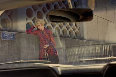 Woman in red patterned dress leaning against a concrete wall, viewed through a car windshield