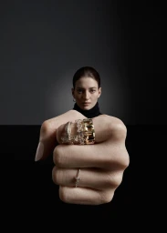 Close-up of a clenched fist displaying multiple gold and diamond rings, with a woman's face blurred in the background.
