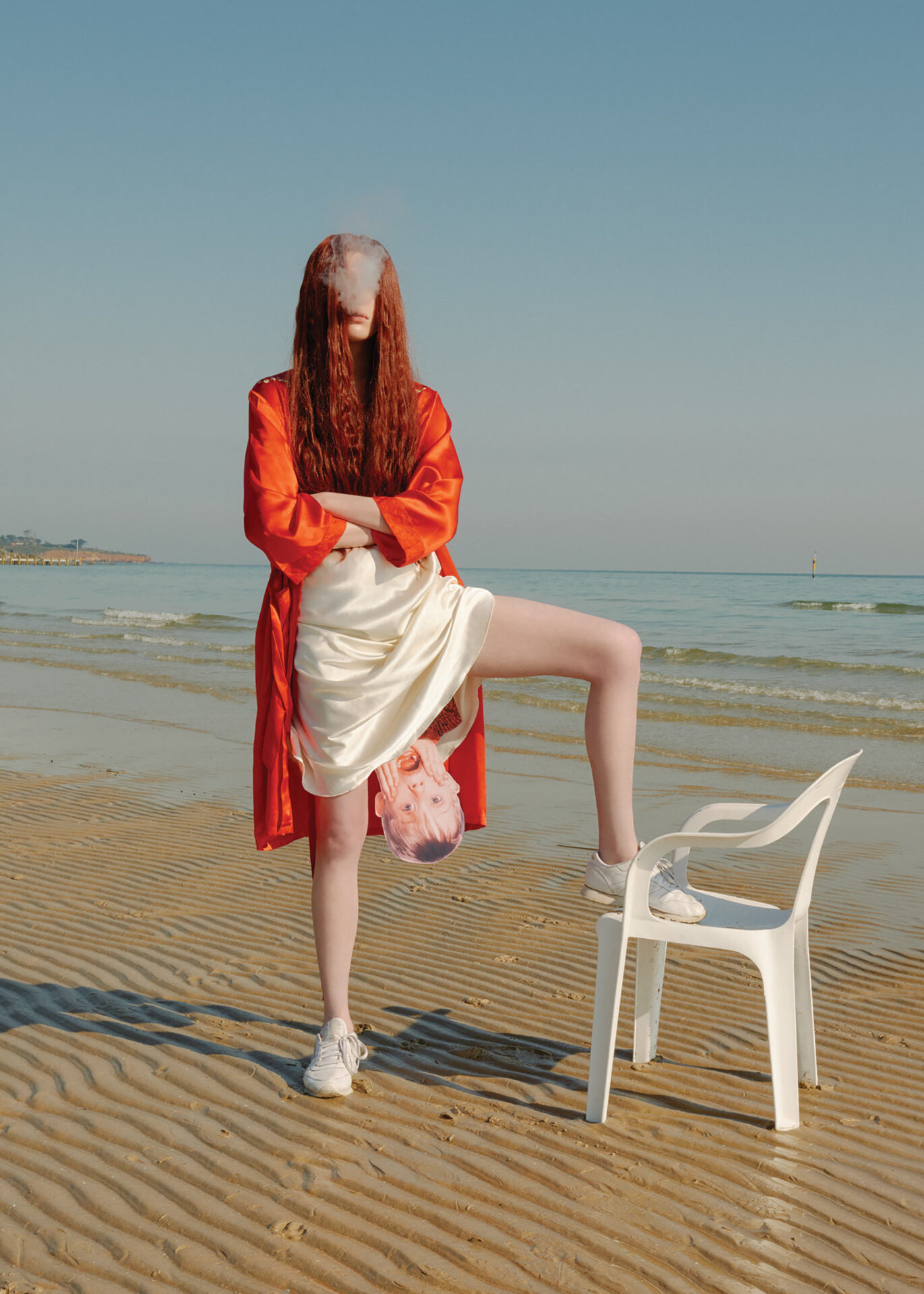 Surreal beach scene features a redhead woman with crossed arms, dressed in orange, while another woman's leg rests on a white chair.