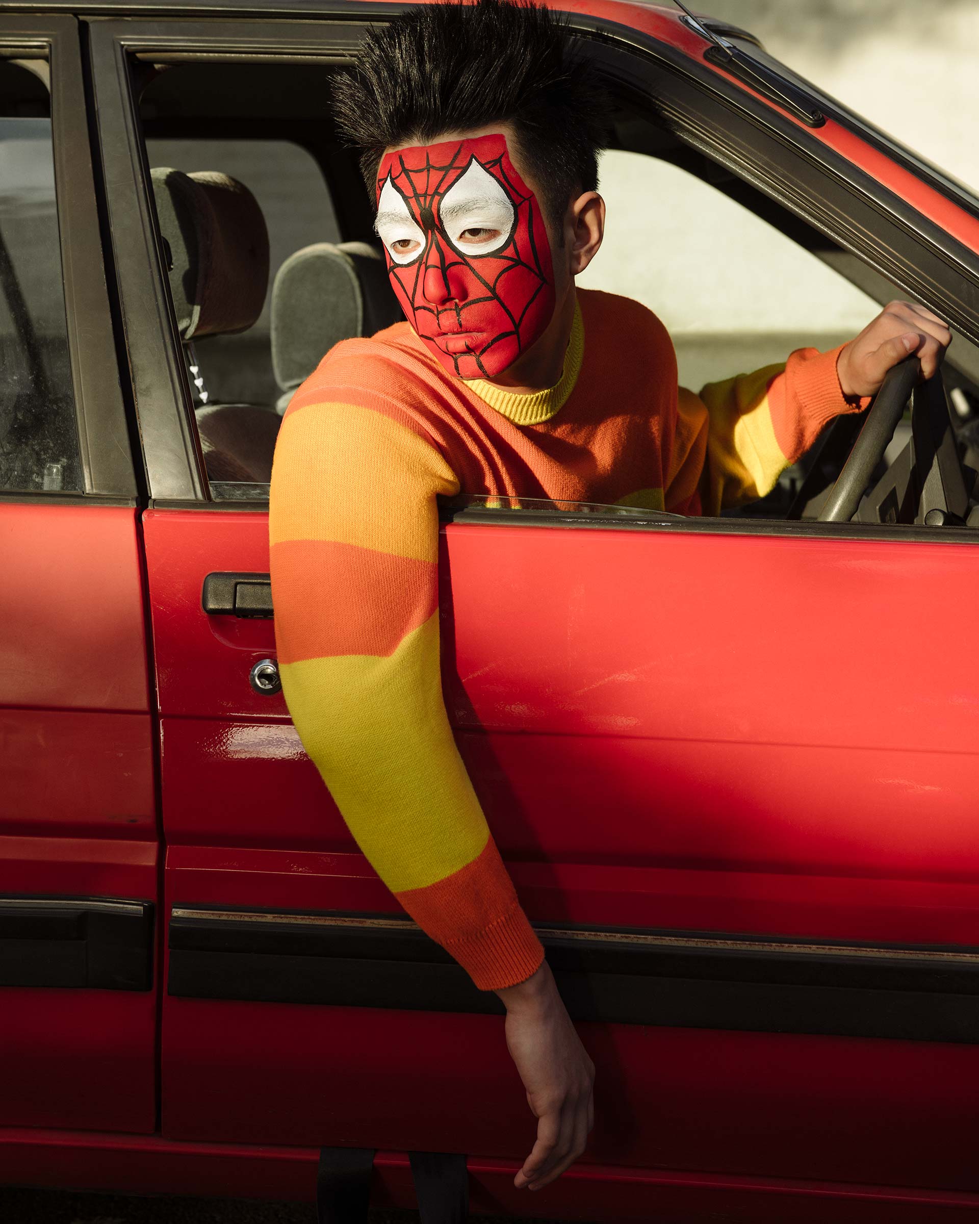 Man with spider web face paint driving a red car, wearing a striped sweater of yellow, orange, and red with his arm out the window.