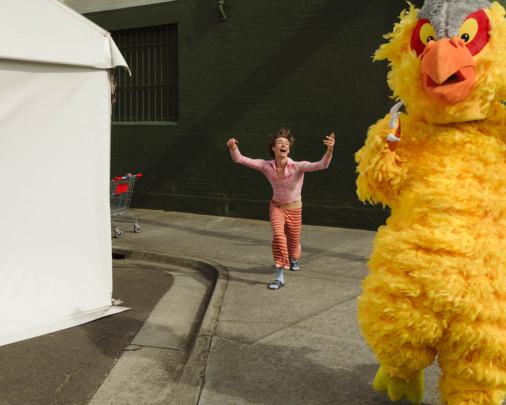 Adult female runs with arms up in the air alongside a large person in a yellow bird mascot costume on an outdoor city street.