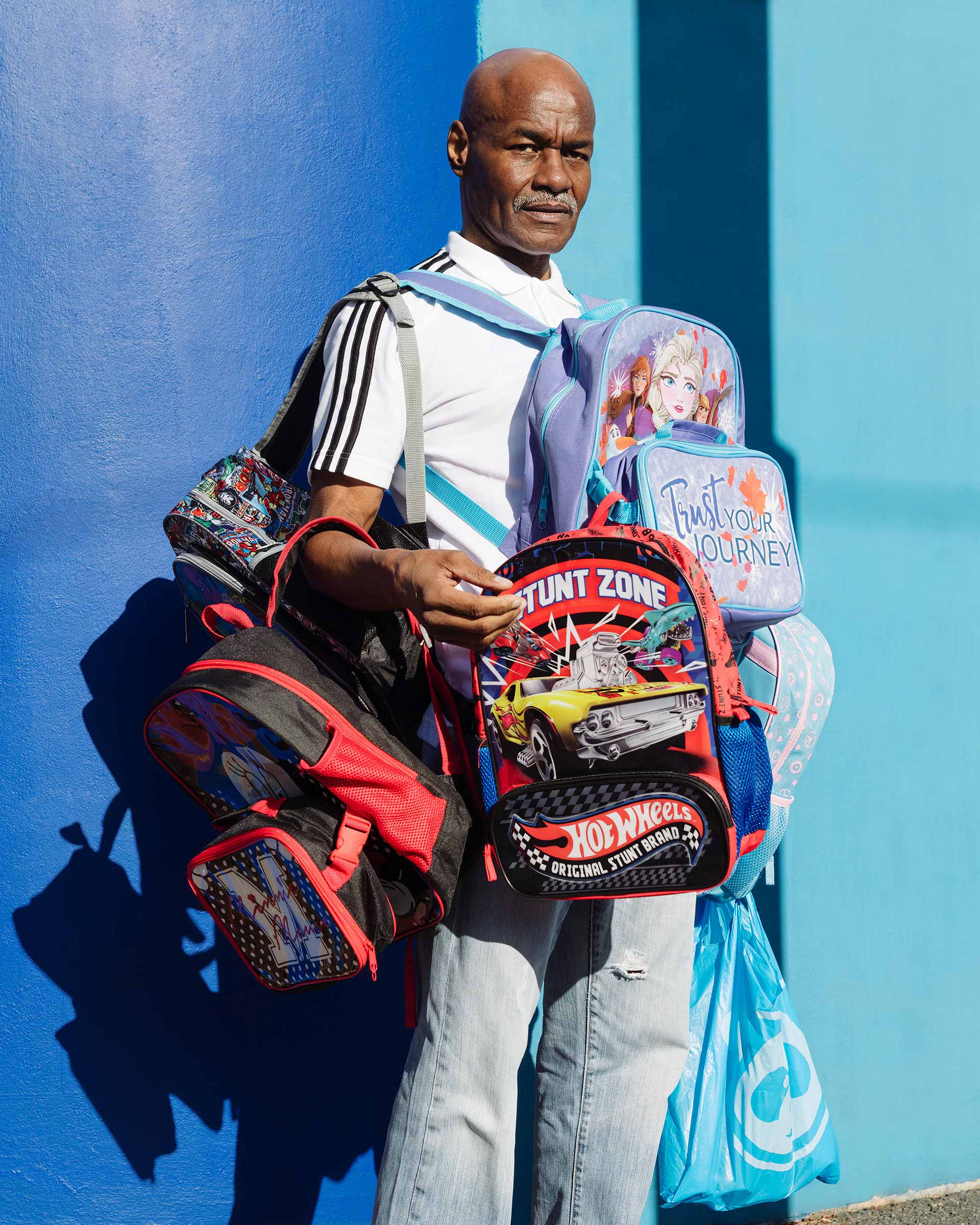 An adult Black man stands outside on a sunny day holding a stack of childrens backpacks and lunchboxes with a blue wall background.