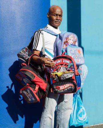 An adult Black man stands outside on a sunny day holding a stack of childrens backpacks and lunchboxes with a blue wall background.