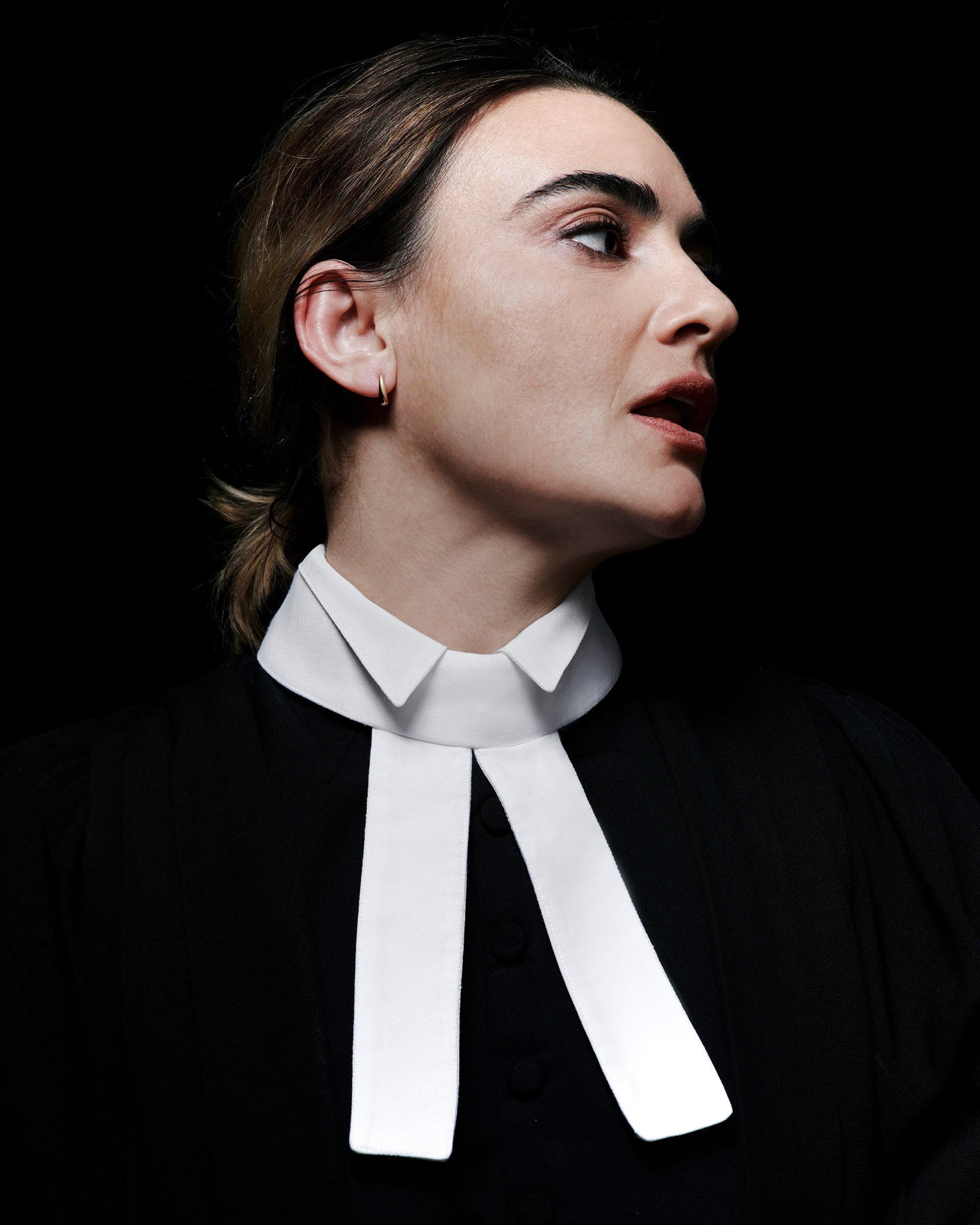 Portrait of adult woman with white collar looking left against black background. Close-up on woman with neutral facial expression.