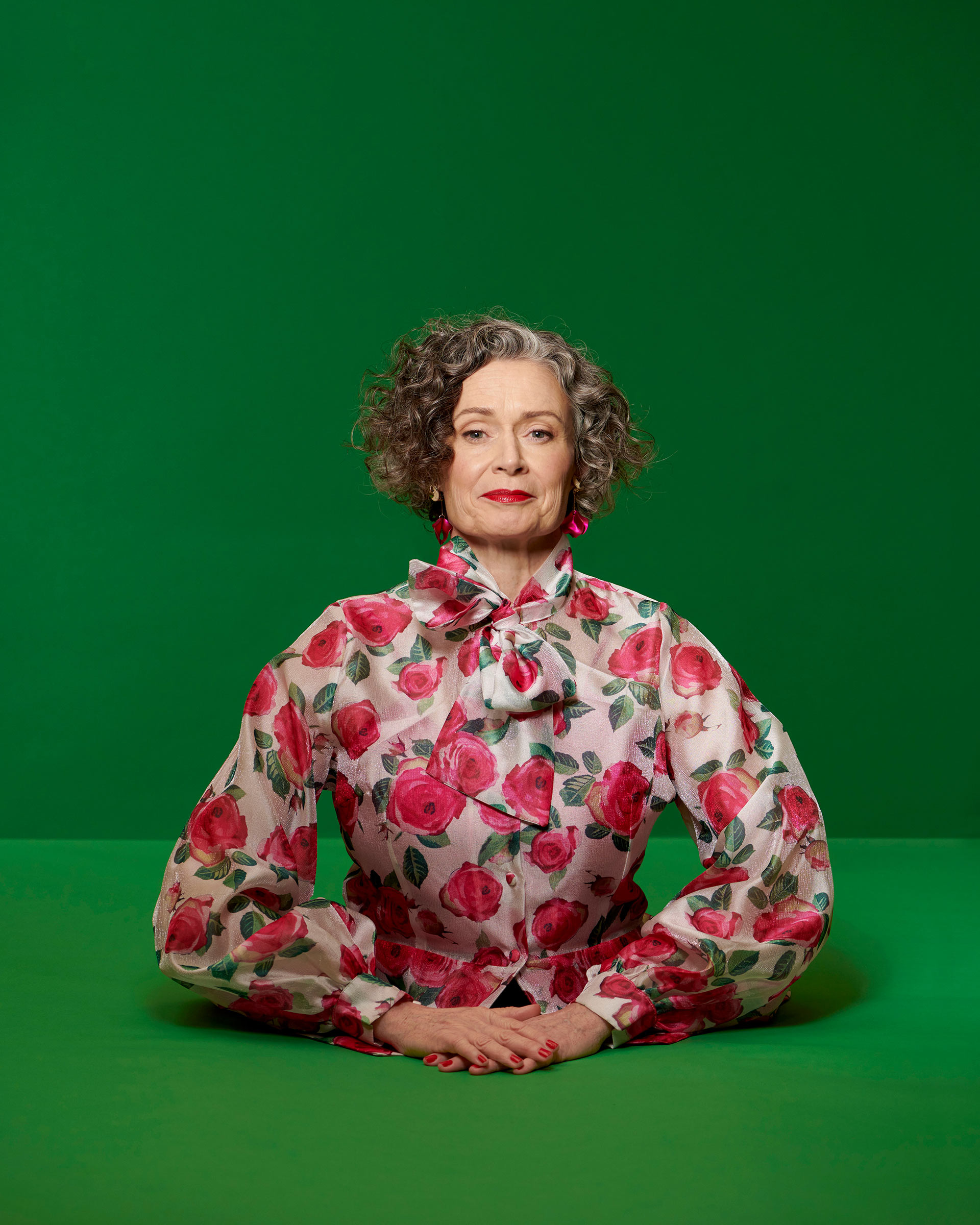 Elegant older woman in a floral blouse sits with hands clasped against a solid green backdrop in a portrait photograph.