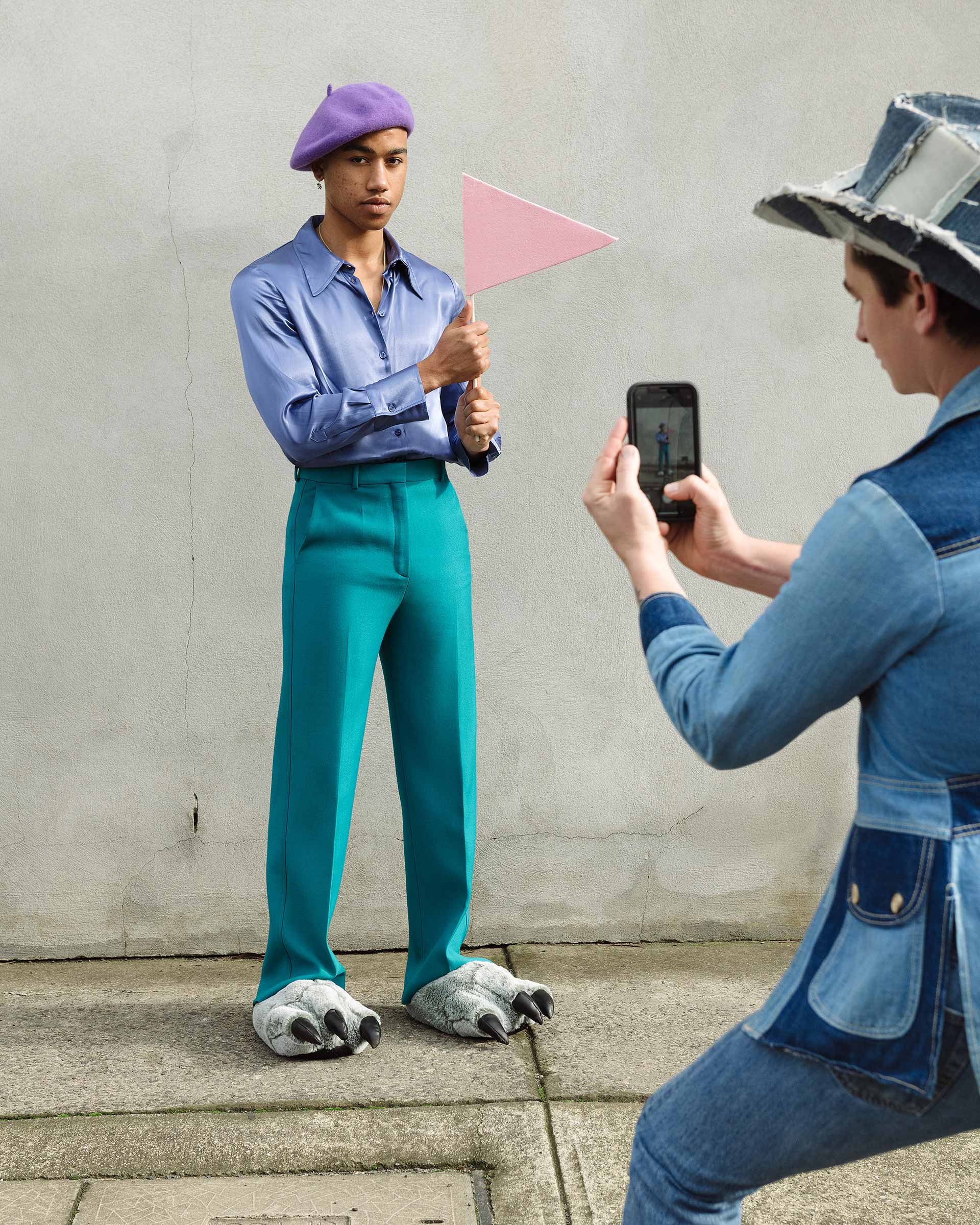 A person in teal pants, animal claw slippers, and a purple hat holds a pink flag as another person photographs them with a smartphone.