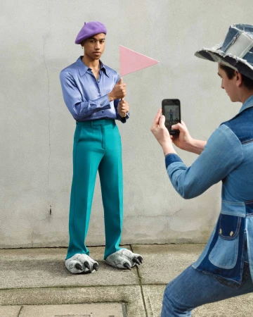 A person in teal pants, animal claw slippers, and a purple hat holds a pink flag as another person photographs them with a smartphone.