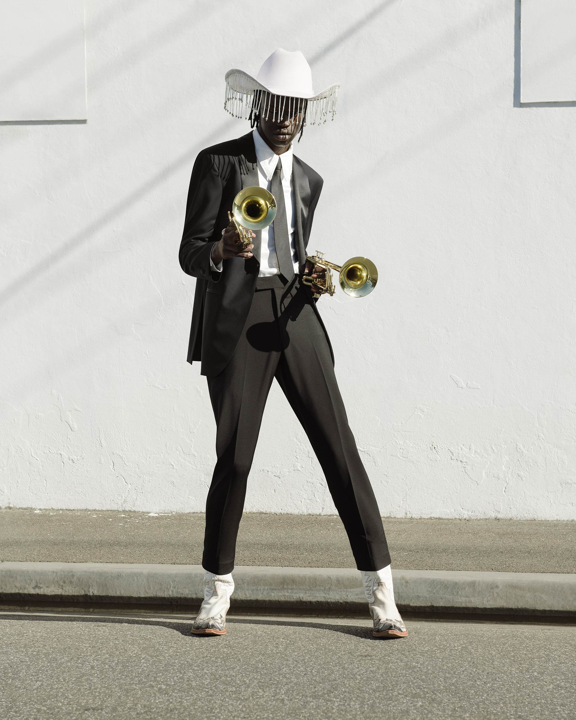 Adult male in a black suit, white cowboy hat with silver fringe, and white cowboy boots, holding two gold trumpets against a white wall.
