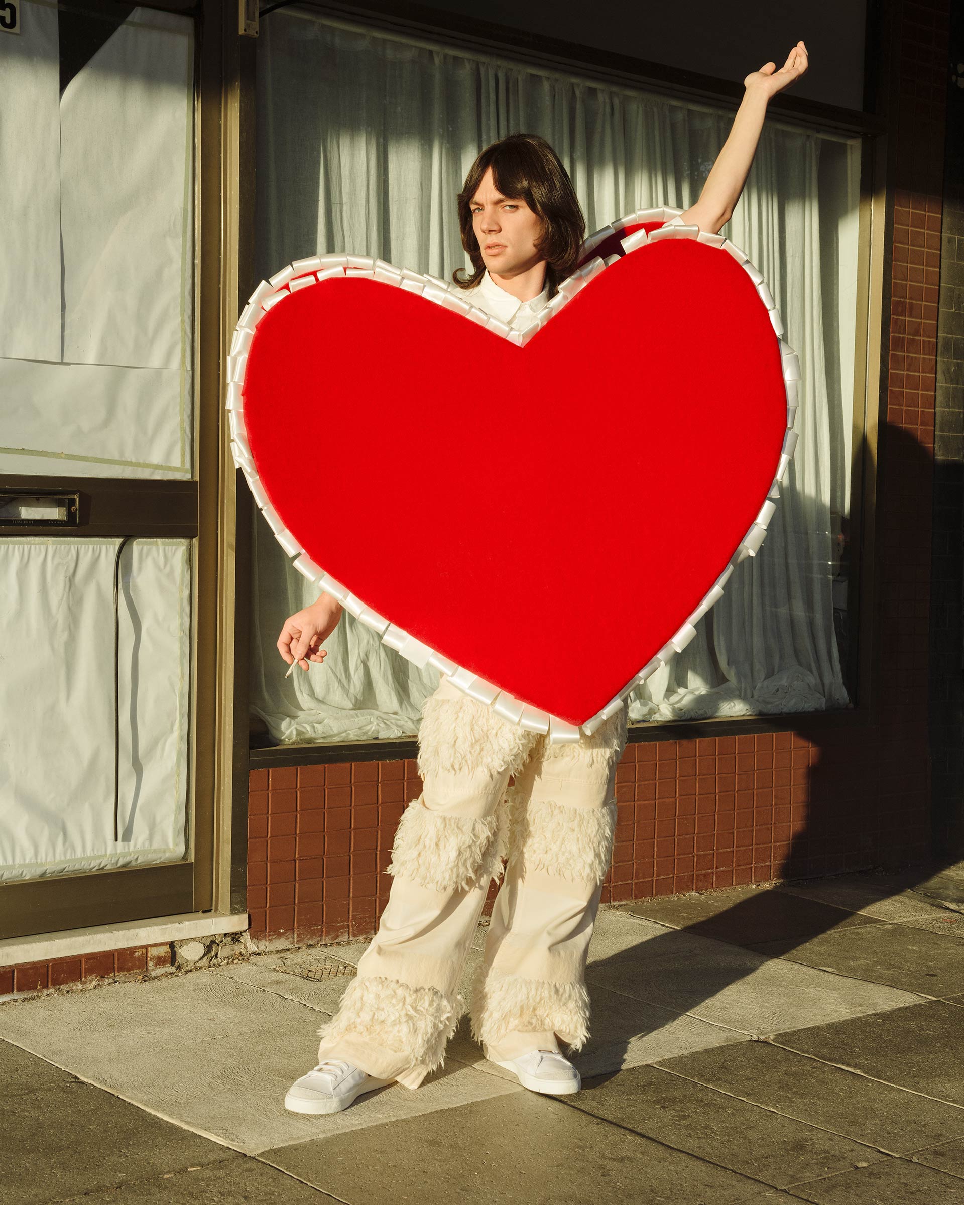 A person stands outside wearing a large red heart costume with fluffy pants, posing in front of a commercial storefront on a sunny day.