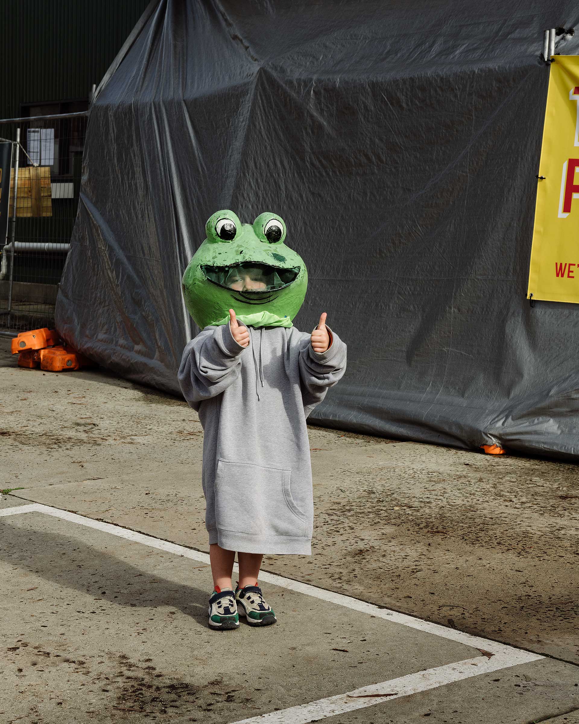 Child wearing a large green frog head and grey hooded sweatshirt gives two thumbs up standing in an outdoor paved area.