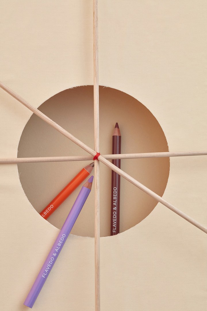Three makeup pencils are arranged with wooden sticks against a neutral backdrop with a circular cut-out creating a geometric composition.