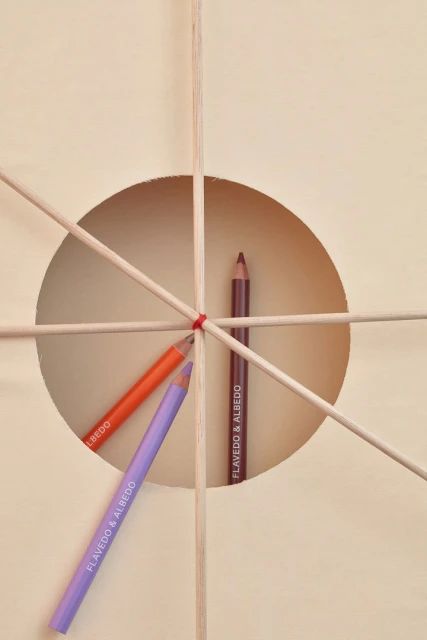 Three makeup pencils are arranged with wooden sticks against a neutral backdrop with a circular cut-out creating a geometric composition.