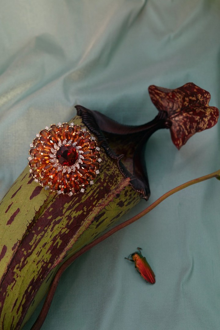 A gemstone brooch sits on a green pitcher plant near an iridescent beetle against a pale blue fabric backdrop in an indoor setting.
