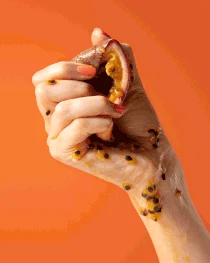 Close-up of a hand squeezing a passion fruit, with seeds and pulp visible against a bright orange background.