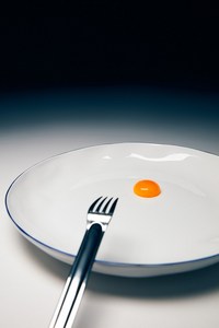 Minimalist food still life: egg yolk on a white plate with a silver fork, set against a dark gradient background. Food photography.
