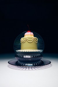 Close-up studio shot of a small, frosted cake with a cherry on top, sitting on stacked glass plates against a dark background.