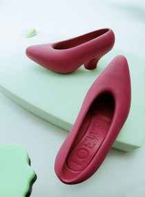 Studio shot of two red Loewe heels. One heel is sitting inside the other, on a light blue platform with some green shapes around.