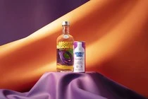 Still life of an Absolut Vodka bottle and a can on a purple silk-draped surface against an orange abstract background.