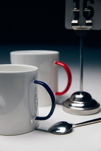 Two white ceramic mugs with blue and red handles sit on a white table beside a spoon and a table number against a dark background.