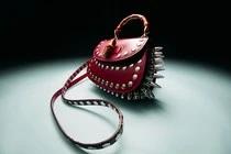 Close-up studio shot of a studded, red leather saddle bag with silver accents and a decorative tassel, against a dark background.