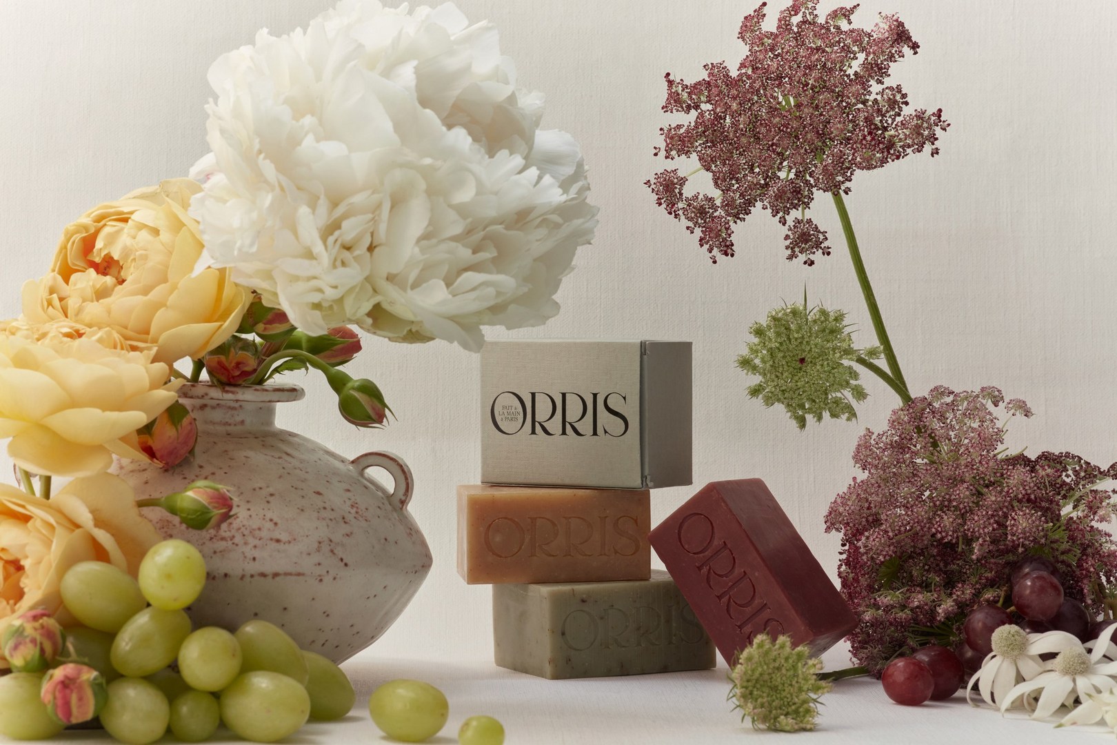 Still life shot of Orris brand soaps stacked amongst floral arrangements and green grapes on a white surface.