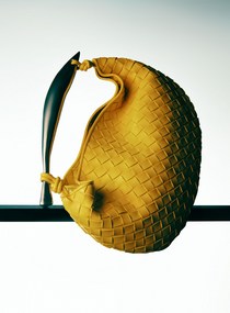 Close-up studio shot of a yellow woven leather handbag with a black handle, showcased on a reflective black surface against a white backdrop.