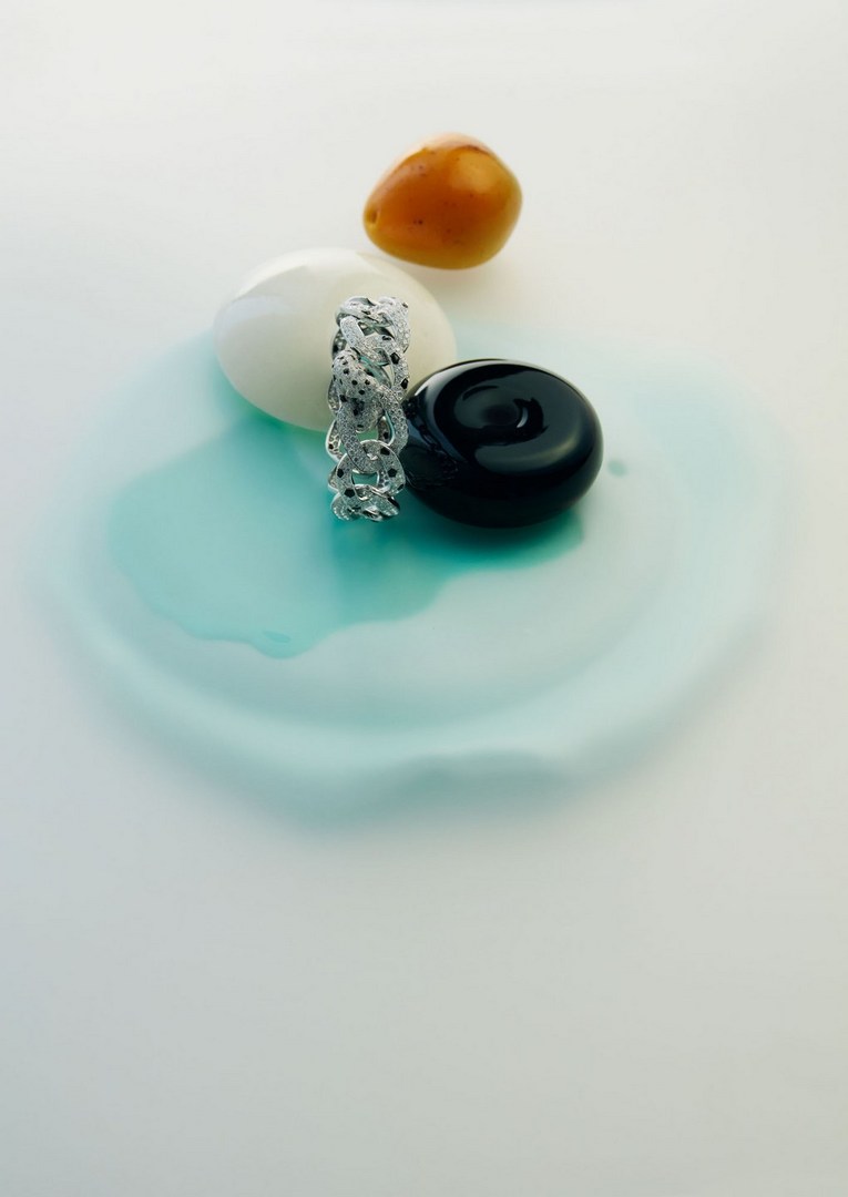 Silver chain link bracelet on a light turquoise surface alongside a white sphere, black disk, and orange cherry tomato.