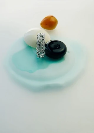 Silver chain link bracelet on a light turquoise surface alongside a white sphere, black disk, and orange cherry tomato.