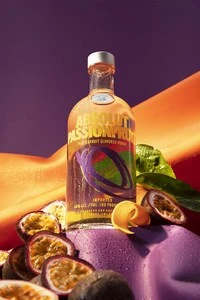 A glass bottle of Absolut Passionfruit vodka surrounded by fresh halved passionfruits on a purple and orange set.