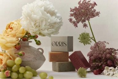 Still life arrangement of Orris soap bars, ceramic vase with white and yellow peonies, grapes, and wildflowers on a pale background.