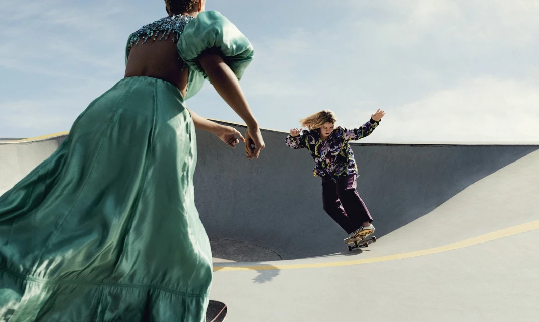 Two people skateboarding in an outdoor skatepark, one in a green dress, the other in a floral shirt and purple pants, under a bright sky.