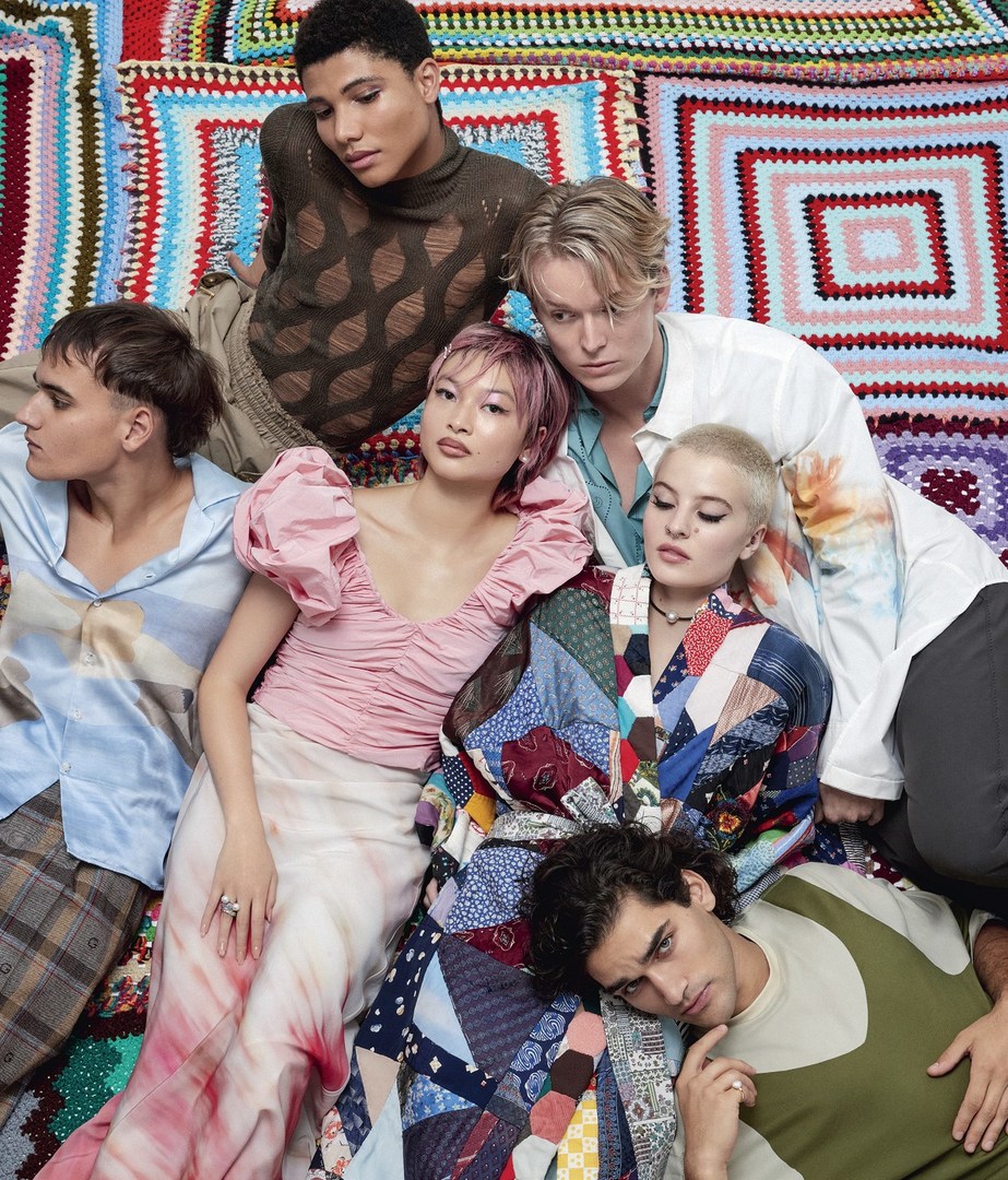 Group portrait of six diverse young adult fashion models in trendy outfits against a colorful knitted patchwork blanket backdrop.