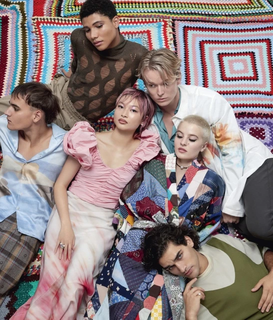 Group portrait of six diverse young adult fashion models in trendy outfits against a colorful knitted patchwork blanket backdrop. 