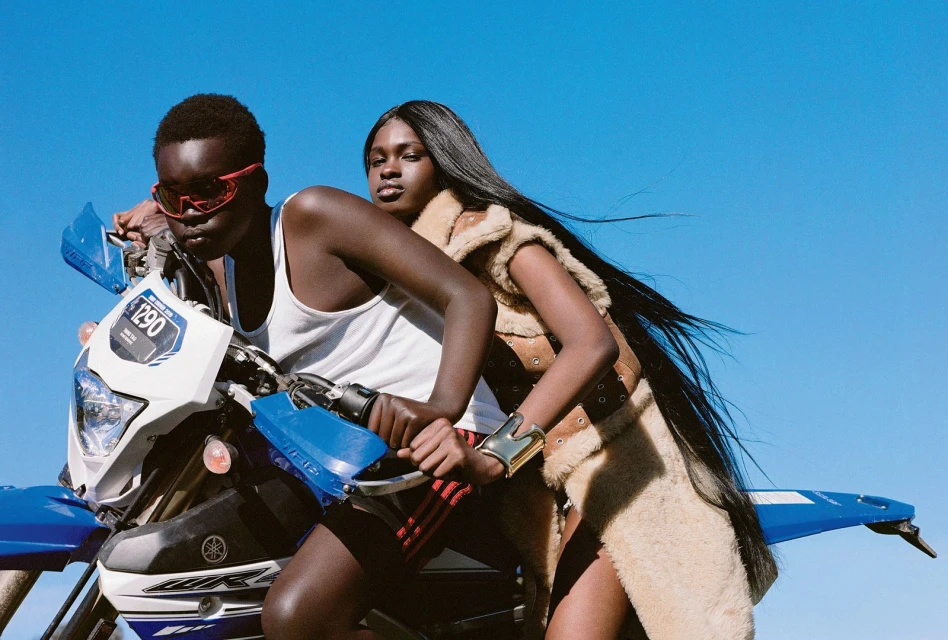 Two dark-skinned women pose on a blue Yamaha dirt bike under a bright blue sky, exuding confidence and style in an outdoor setting.