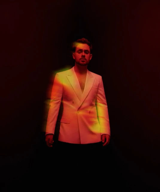 Man in cream suit, posing in front of a black background with a prism effect in studio lighting. Confident gaze, formal attire.